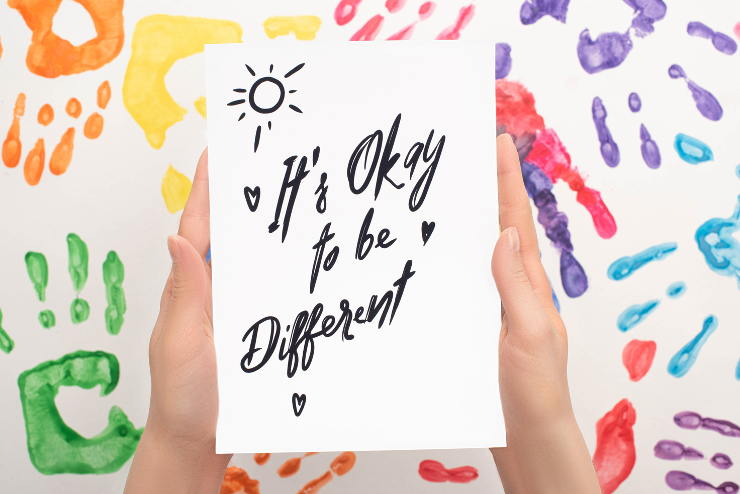 cropped view of woman holding card with Its Okay to be different lettering on white with hand prints